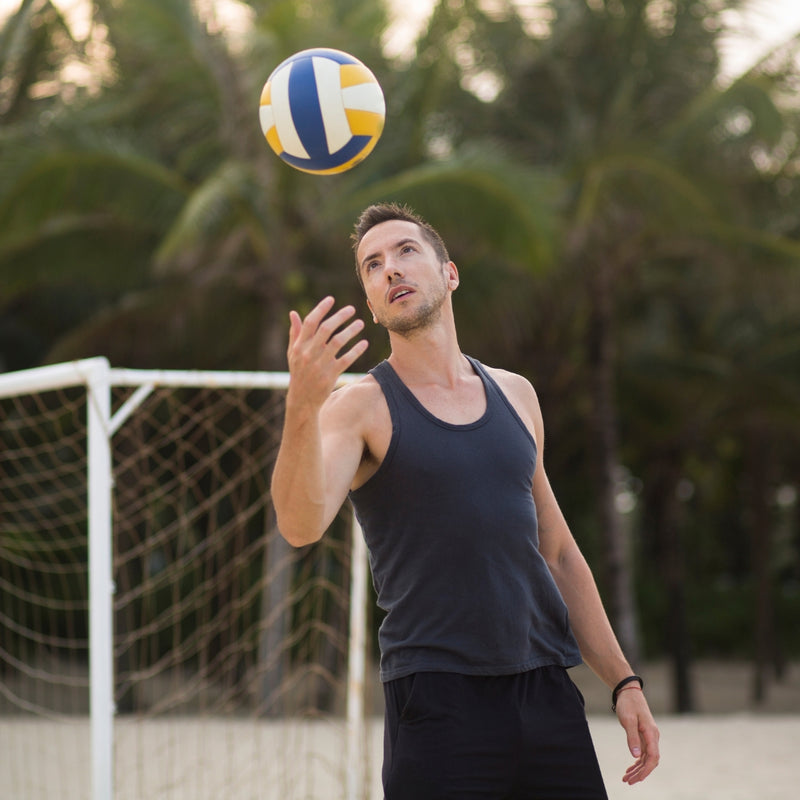 A man in a dark navy tank top and black shorts on a beach volleyball court, looking up at a yellow, white, and blue volleyball. Palm trees and a volleyball goal are visible in the background.