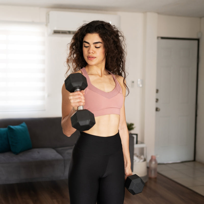 A woman with curly dark hair stands in a living room setting, holding a black hexagonal dumbbell in each hand. She is wearing a dusty pink sports bra and black leggings.