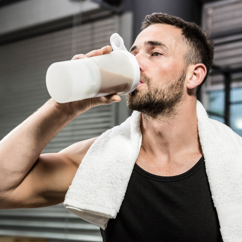 Man drinking from a white protein shake container with a towel around his neck.