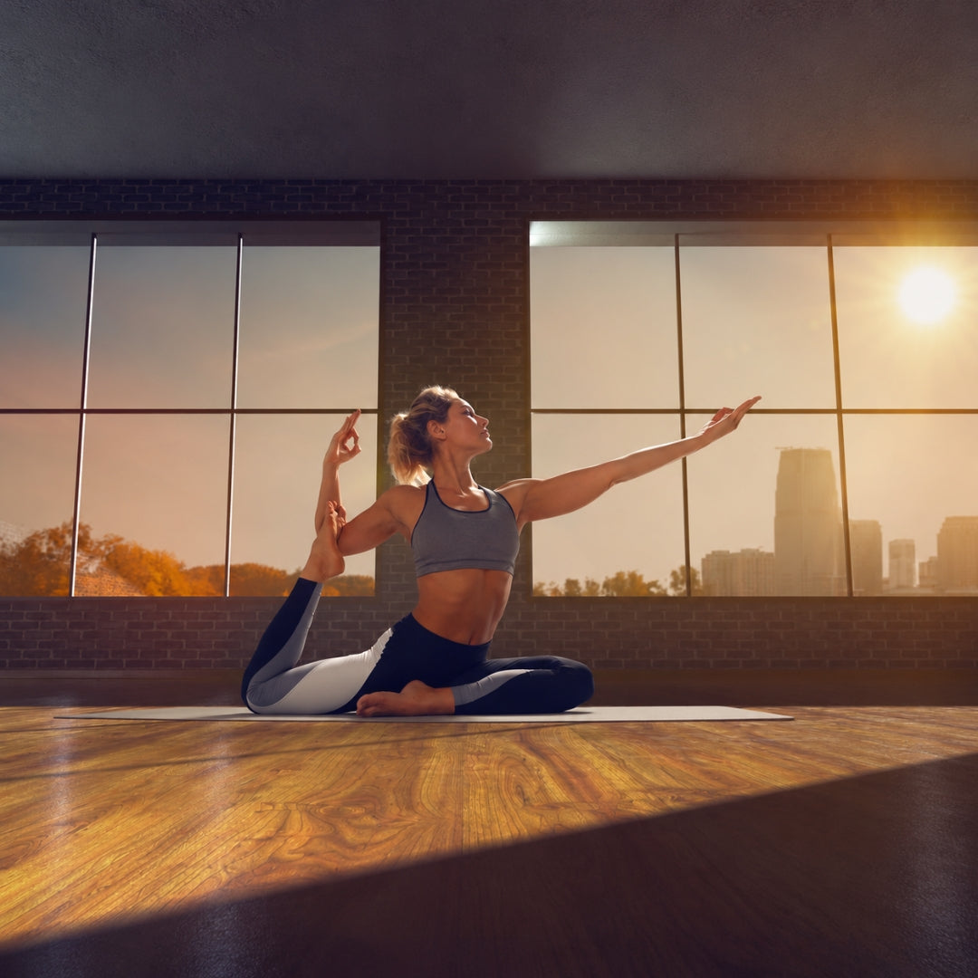 A woman in a gray sports bra and black leggings with white side panels performs a yoga pose on a mat inside a sunlit studio with large windows and a city skyline view.